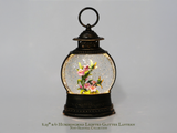A 3.25" b/o Hummingbird Lighted Glitter Lantern from the Non-Seasonal Collection, featuring a dark bronze metal frame with a clear glitter-filled globe containing a pink and yellow hummingbird flower design, set against a neutral gray background.