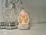 A small decorative lantern-shaped snow globe with a glowing angel figure inside, placed on a tabletop next to a clear crystal ornament and metallic decorative pieces against a soft neutral background.
