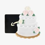 A white two-tiered wedding cake decoration or figurine with pink and white roses, green leaves, and a scalloped gold base, shown in front of a black square box with a gold arrow logo on a light gray background.