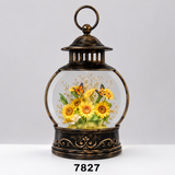 A decorative lantern-shaped glass display with an antique bronze metal frame and base, containing a preserved floral arrangement of yellow sunflowers, green leaves, and orange-and-black butterflies under clear glass. The lantern is centered on a light gray studio background with a white surface, and the number 7827 appears below.