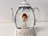 A white ceramic teapot-shaped snow globe figurine featuring a small angel figure inside a clear glass dome with silver glitter, set on an ornate white base against a plain light background.
