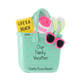 Personalized beach-themed bucket with a mint green polka dot exterior, filled with summer accessories including a yellow 'LIFE'S A BEACH' sign, pink and white striped towel, flip-flops, and pink sunglasses. The bucket is printed with 'Our Family Vacation' and 'Santa Rosa Beach'.