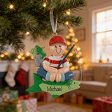 A personalized ornament hanging from a Christmas tree, featuring a smiling boy in a red hat and striped shirt, holding a fishing rod and accompanied by a fish.