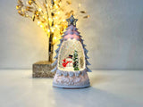 A Christmas tree-shaped snow globe figurine with a glittery white interior, a snowman and small tree scene, and a silver star on top, displayed on a white surface with a softly lit decorative tree in the background.