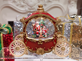 A red and gold ornate carriage-shaped Christmas ornament with Santa and a tree inside a clear snow globe, displayed on a clear stand with a sign reading 'Red Carriage Christmas Collection.'.