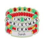 A personalized Christmas-themed plaque or ornament featuring stacked beaded bracelets in red, green, and yellow, with white letter tiles spelling 'year IN MY CHRISTMAS ERA' and a white banner at the bottom personalized with the name 'Sarah'.