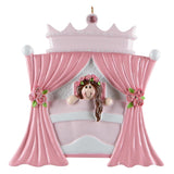 A pink princess-themed ornament shaped like a stage with curtains, a crown, and a small girl peeking out from behind, shown on a plain white background.