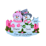 Personalized Christmas snowman ornament featuring two snowmen named Sherri and David with pink and green scarves, pink hats, a pink fence, green holly accents, and a blue base that reads 'Our First Christmas' and 'year'.