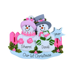 Personalized Christmas snowman ornament featuring two snowmen named Sherri and David with pink and green scarves, pink hats, a pink fence, green holly accents, and a blue base that reads 'Our First Christmas' and 'year'.