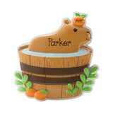A custom capybara figurine sitting in a wooden barrel tub, decorated with the name 'Parker' on its snout, an orange 'year' on its head, and green leaves and oranges at the base, shown on a white background.