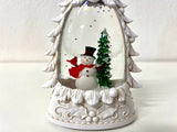 A white ceramic snow globe figurine featuring a snowman with a red scarf and black top hat next to a small green Christmas tree, set against a white background.