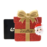 A personalized Christmas gift box ornament in red glitter with a gold bow and ribbon, featuring the name 'Jonathan' on the front and a hanging Santa tag marked '2026'. A black gift box is partially visible behind it on a white background.