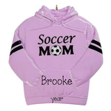 A glossy lavender purple Christmas ornament shaped like a hoodie with a gold hanging loop at the hood. The front features the text 'Soccer MOM' in black with a soccer ball graphic, 'Brooke' in script, and 'year' at the bottom.